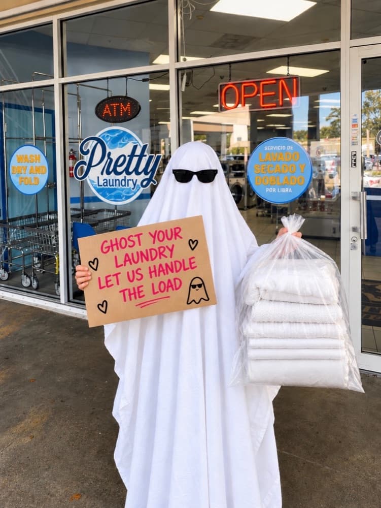 Person in a ghost costume holding a sign that reads Ghost your laundry, let us handle the load, outside Pretty Laundry